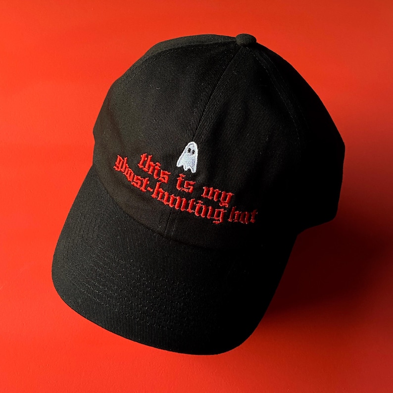 Op de afbeelding: Zwarte baseballpet met een witte geest en rode geborduurde tekst die "This is my ghost-hunting hat." zegt.