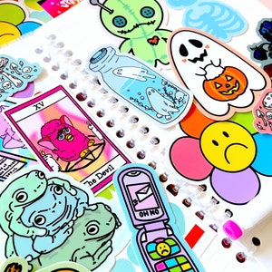 May include: A collection of colorful stickers featuring a variety of designs, including a ghost holding a jack-o-lantern, a fuzzy pink creature, a frog, a phone with a sad face, and a tarot card with the text "The Devil" and the number "XV".