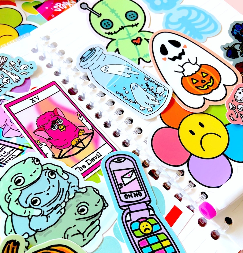 May include: A collection of colorful stickers featuring a variety of designs, including a ghost holding a jack-o-lantern, a fuzzy pink creature, a frog, a phone with a sad face, and a tarot card with the text "The Devil" and the number "XV".