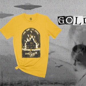 Op de afbeelding: Een geel T-shirt met een zwart-witte afbeelding van een brandend huis met een skelet dat ervoor ligt. De tekst "The End Is Here" staat in een cirkel boven het huis. Het woord "GOLD" staat in zwart-witte letters op een witte achtergrond.