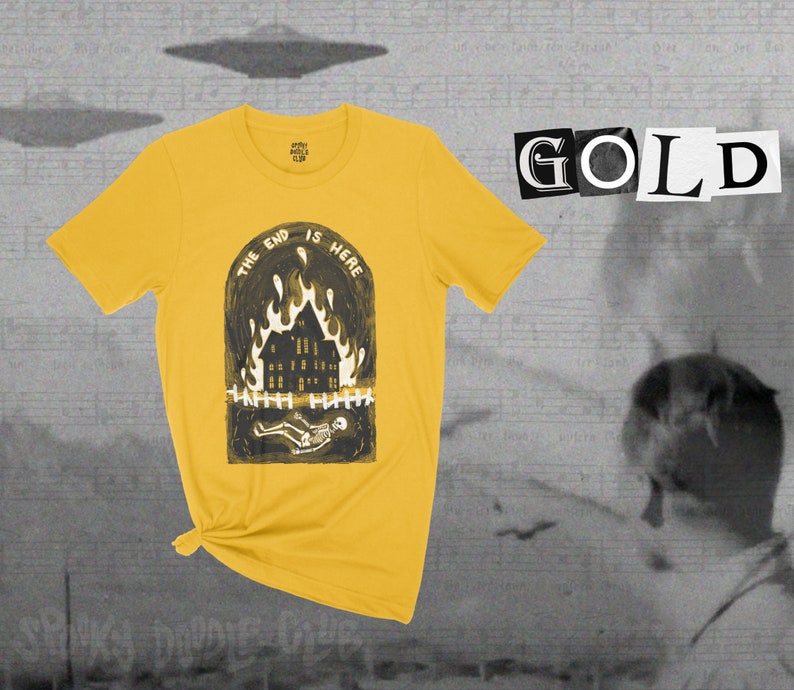 Op de afbeelding: Een geel T-shirt met een zwart-witte afbeelding van een brandend huis met een skelet dat ervoor ligt. De tekst "The End Is Here" staat in een cirkel boven het huis. Het woord "GOLD" staat in zwart-witte letters op een witte achtergrond.