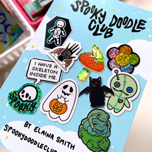 May include: A blue card with the text "Spooky Doodle Club" and a variety of colorful, glittery stickers featuring Halloween themes, including a skeleton, a ghost, a cat, a frog, a flower, and a voodoo doll. The text "By Elaina Smith Spookydoodleclub.etsy.com" is at the bottom of the card.