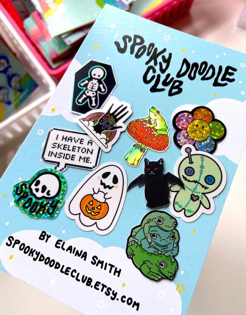 May include: A blue card with the text "Spooky Doodle Club" and a variety of colorful, glittery stickers featuring Halloween themes, including a skeleton, a ghost, a cat, a frog, a flower, and a voodoo doll. The text "By Elaina Smith Spookydoodleclub.etsy.com" is at the bottom of the card.