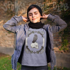 May include: A light blue t-shirt featuring a raven perched on a skull, surrounded by candles and skeleton hands. The design is framed by a green vine. The words "Spooky Doodle Club" are at the top.