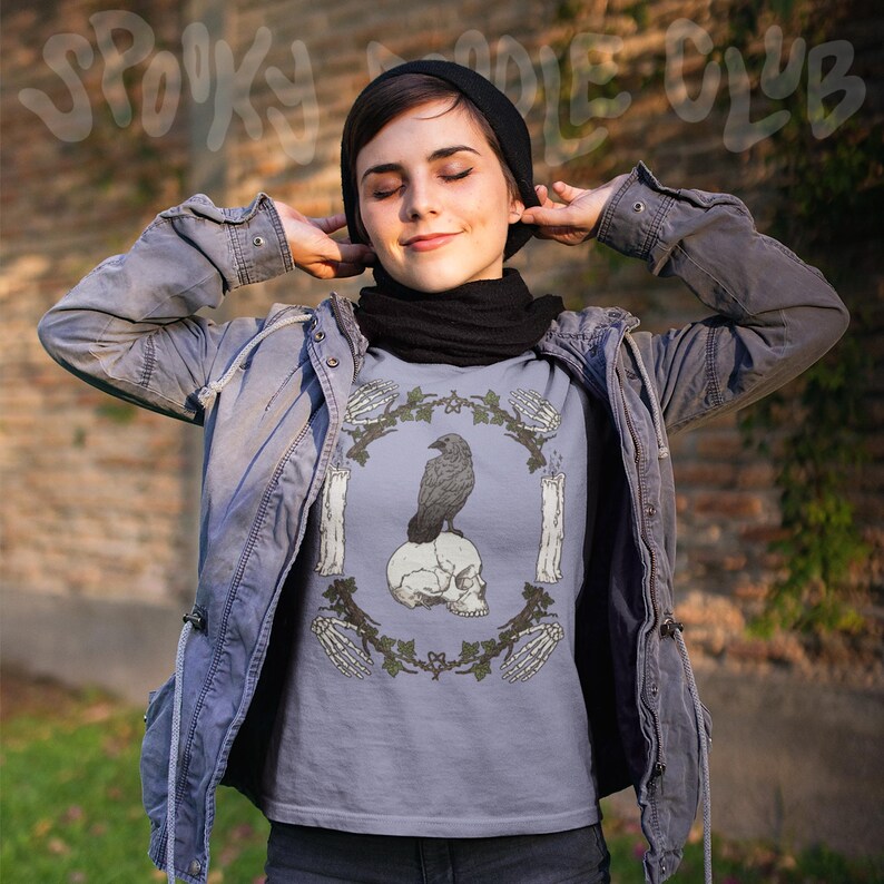 May include: A light blue t-shirt featuring a raven perched on a skull, surrounded by candles and skeleton hands. The design is framed by a green vine. The words "Spooky Doodle Club" are at the top.