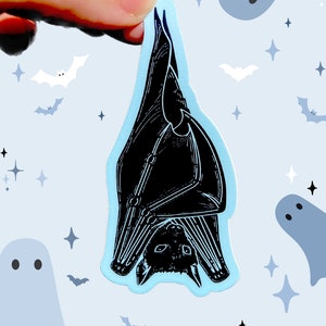 May include: A black bat sticker with a blue background. The bat is hanging upside down with its wings spread out. The sticker is surrounded by white stars and ghosts.