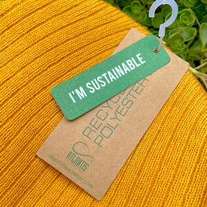 May include: A yellow knitted beanie with a brown tag that says "I'm Sustainable" and "Recycled Polyester" with the brand name "Atlantis" and website address atlantisheadwear.com.