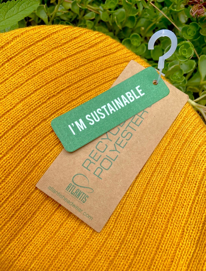 May include: A yellow knitted beanie with a brown tag that says "I'm Sustainable" and "Recycled Polyester" with the brand name "Atlantis" and website address atlantisheadwear.com.