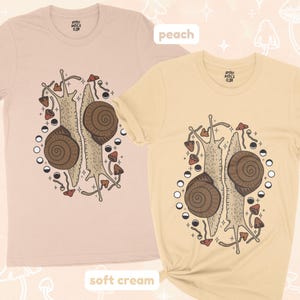 Pu&ograve; includere: Due t-shirt con un design grafico di due lumache con funghi e fasi lunari. Le t-shirt sono nei colori crema morbido e pesca.