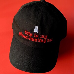 Op de afbeelding: Zwarte baseballpet met rode geborduurde tekst die "This is my ghost-hunting hat" zegt en een klein wit spook.