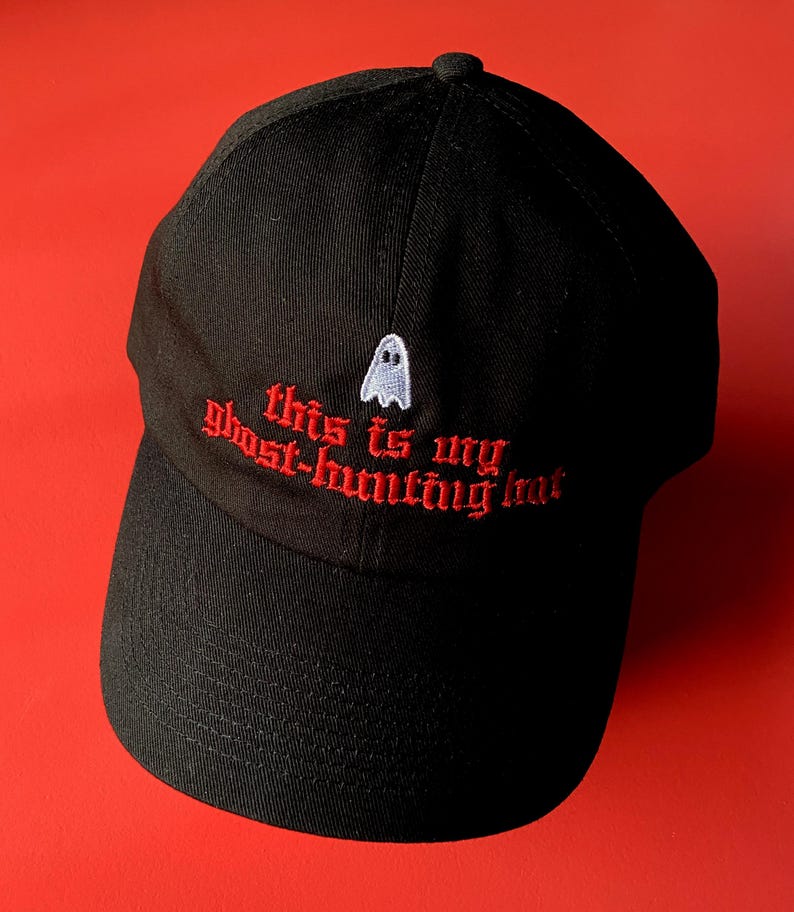 Op de afbeelding: Zwarte baseballpet met rode geborduurde tekst die "This is my ghost-hunting hat" zegt en een klein wit spook.
