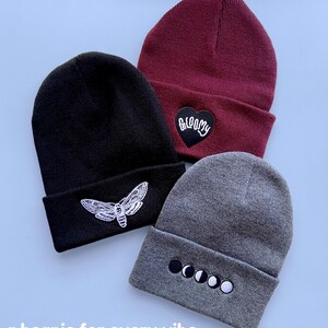 May include: Three knit beanies with different embroidered designs. The beanie on the left is black with a white moth design. The beanie in the middle is burgundy with a black heart that says "GLOOMY" in white. The beanie on the right is gray with a white moon phase design.