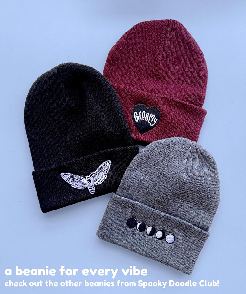 May include: Three knit beanies with different embroidered designs. The beanie on the left is black with a white moth design. The beanie in the middle is burgundy with a black heart that says "GLOOMY" in white. The beanie on the right is gray with a white moon phase design.
