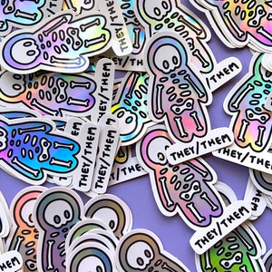 May include: A collection of iridescent holographic stickers featuring a cartoon skeleton figure with the text "They/Them" on each sticker.