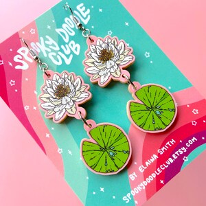 Op de afbeelding: Hangende oorbellen met een waterlelie-ontwerp. De oorbellen hebben een witte en gele bloem, een groen waterlelieblad en roze accenten. De oorbellen hebben zilverkleurige haken. De achtergrond is een kaart met de tekst "Spooky Doodle Club".