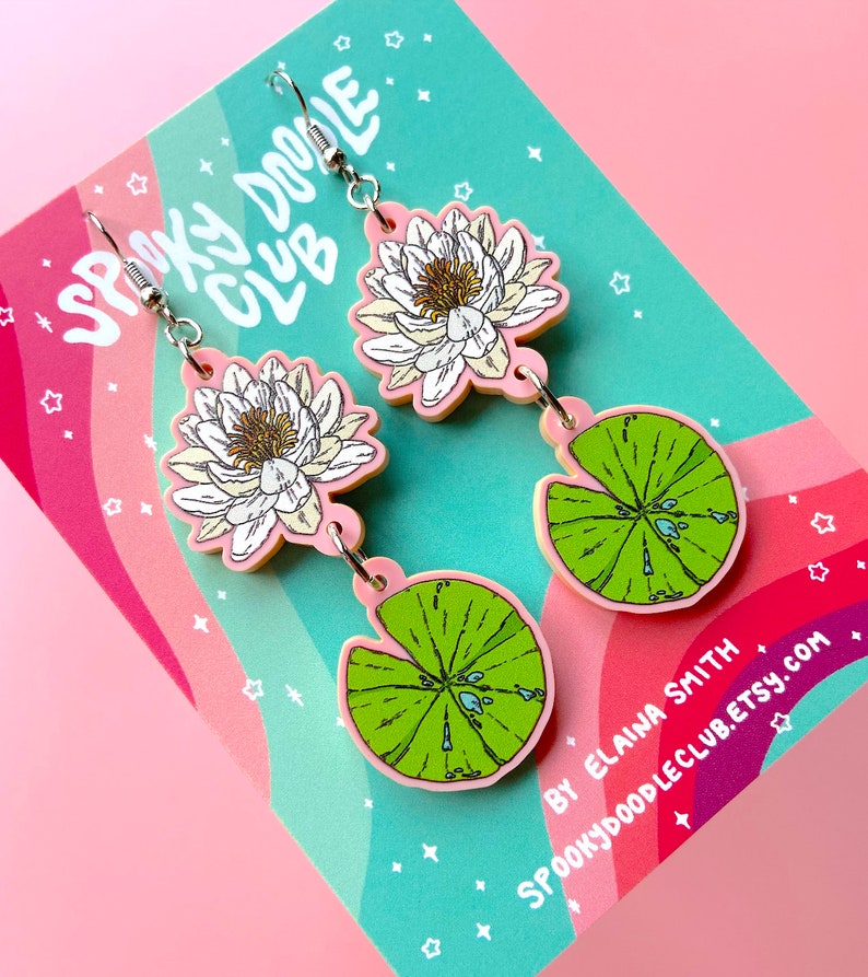 Op de afbeelding: Hangende oorbellen met een waterlelie-ontwerp. De oorbellen hebben een witte en gele bloem, een groen waterlelieblad en roze accenten. De oorbellen hebben zilverkleurige haken. De achtergrond is een kaart met de tekst "Spooky Doodle Club".