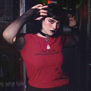 Op de afbeelding: Een vrouw draagt een rood T-shirt met de tekst "This is my ghost-hunting shirt" en een kleine geestenafbeelding. Ze draagt zwarte fishnet handschoenen en een zilveren choker met een geestenbedel.