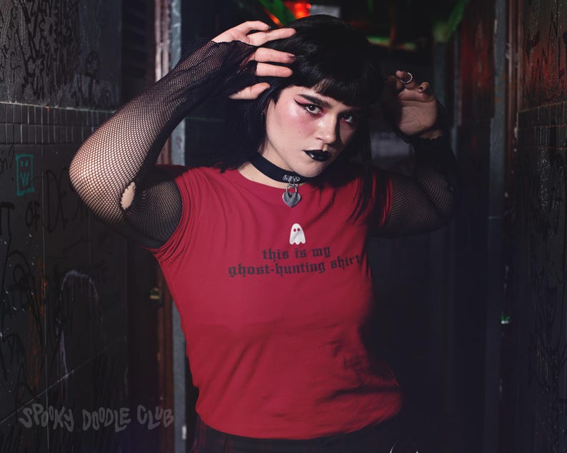 Op de afbeelding: Een vrouw draagt een rood T-shirt met de tekst "This is my ghost-hunting shirt" en een kleine geestenafbeelding. Ze draagt zwarte fishnet handschoenen en een zilveren choker met een geestenbedel.