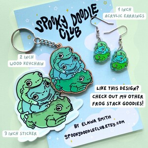 May include: A set of frog stack themed goods, including a 3 inch sticker, a 2 inch wooden keychain, and a pair of 1 inch acrylic earrings. The sticker and keychain feature a stack of three frogs in shades of green and blue. The earrings are also a stack of three frogs, but are smaller and made of clear acrylic. The text "Spooky Doodle Club" is visible in the image.