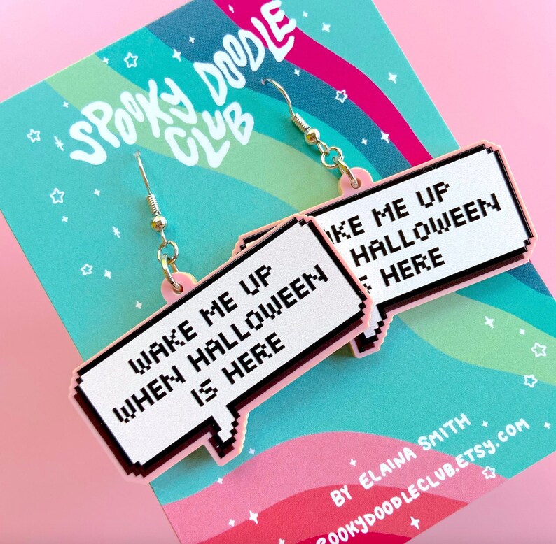 May include: Pink and white statement earrings with a pixelated speech bubble design. The text on the earrings reads "Wake me up when Halloween is here".