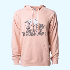 Pode incluir: Um moletom rosa claro com cord&atilde;o branco e bolso canguru. Apresenta um gr&aacute;fico em preto e branco de um rato gritando e o texto "LIFE IS SCARY AND IT'S OK TO SCREAM ABOUT IT".