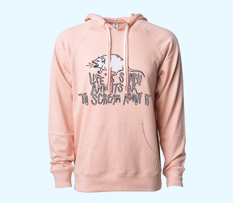 Pode incluir: Um moletom rosa claro com cord&atilde;o branco e bolso canguru. Apresenta um gr&aacute;fico em preto e branco de um rato gritando e o texto "LIFE IS SCARY AND IT'S OK TO SCREAM ABOUT IT".