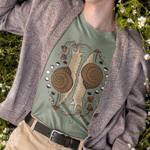 Pu&ograve; includere: Una maglietta verde con una grafica di due lumache con disegni di funghi e lune. La maglietta &egrave; indossata da una persona con un cardigan marrone.