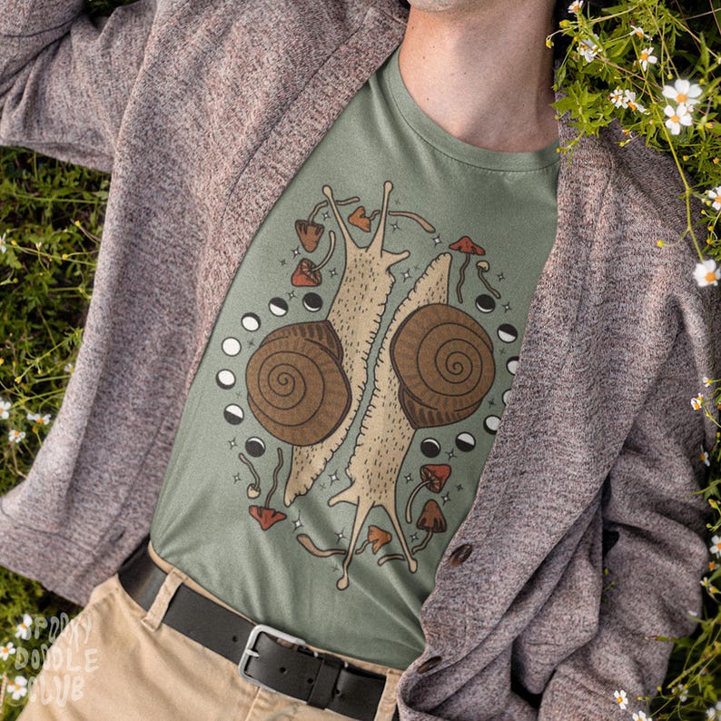Pu&ograve; includere: Una maglietta verde con una grafica di due lumache con disegni di funghi e lune. La maglietta &egrave; indossata da una persona con un cardigan marrone.