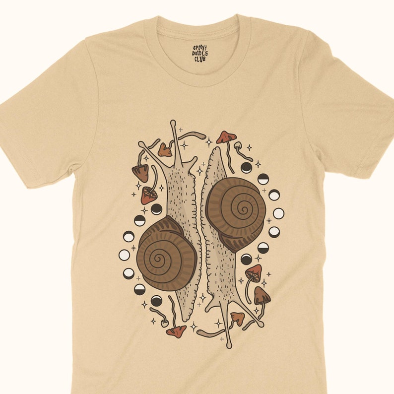 Pu&ograve; includere: Una maglietta beige con un design grafico di due lumache con i loro gusci uno di fronte all'altro. Le lumache sono circondate da funghi e lune crescenti. Il testo "Sleepy Doodle Club" &egrave; stampato sulla maglietta.