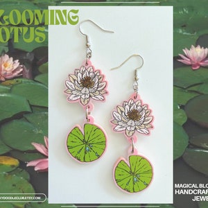 Op de afbeelding: Roze en witte lotusbloem oorbellen met groene waterlelies. De oorbellen zijn gemaakt van acryl en hebben een gedetailleerd bloemenontwerp. De tekst "BLOOMING LOTUS" staat in het groen bovenaan. De tekst "MAGICAL BLOOMS HANDCRAFTED JEWELRY" staat onderaan.
