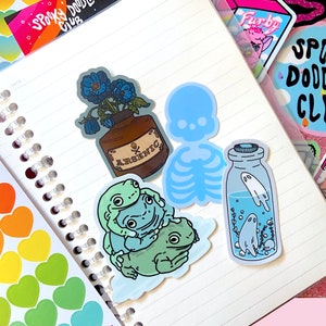 May include: A set of five stickers featuring a blue skeleton, a bottle with a ghost inside, three frogs, and a bottle with blue flowers and the word "ARSENIC" on the label.