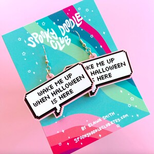 May include: Pink and white statement earrings with the text "Wake me up when Halloween is here" in a pixelated font. The earrings are shaped like speech bubbles.