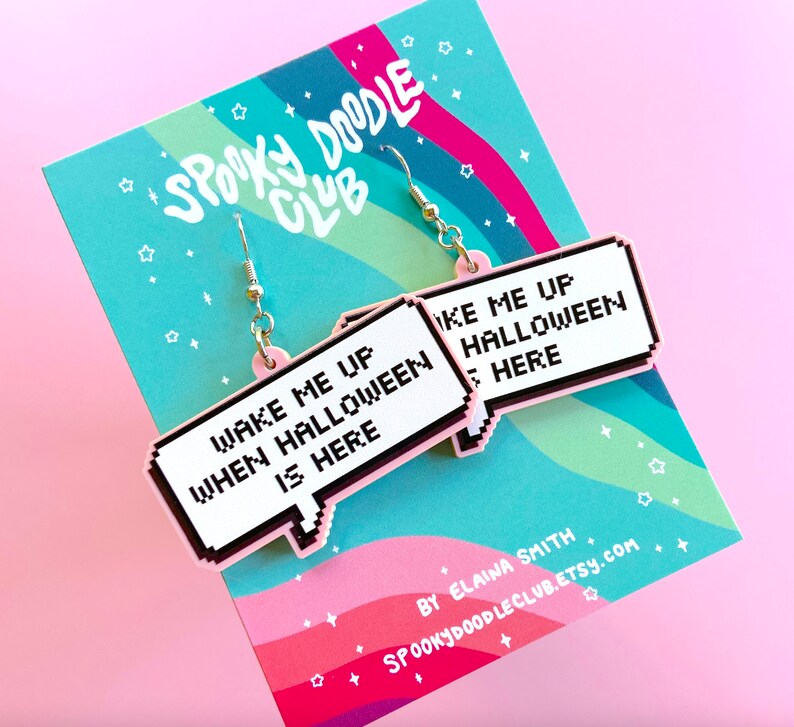 May include: Pink and white statement earrings with the text "Wake me up when Halloween is here" in a pixelated font. The earrings are shaped like speech bubbles.