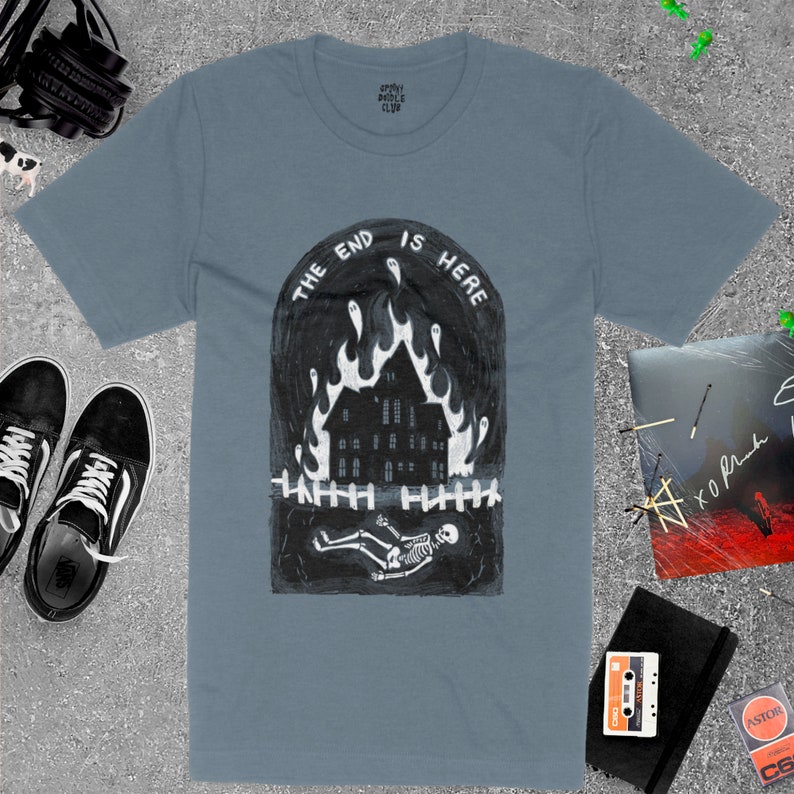Op de afbeelding: Een lichtblauw t-shirt met een zwart-witte illustratie van een brandend huis met een skelet dat ervoor ligt. De tekst "THE END IS HERE" staat boven het huis.