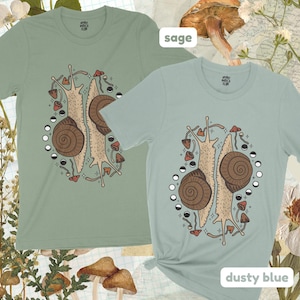 Pu&ograve; includere: Due t-shirt di colore verde salvia e blu polvere. Entrambe le magliette presentano una grafica di due lumache con funghi e lune. Il testo "Sage" e "Dusty Blue" &egrave; stampato sul fondo delle magliette.