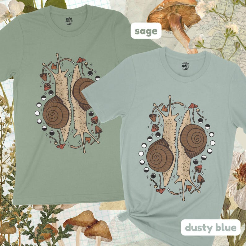 Pu&ograve; includere: Due t-shirt di colore verde salvia e blu polvere. Entrambe le magliette presentano una grafica di due lumache con funghi e lune. Il testo "Sage" e "Dusty Blue" &egrave; stampato sul fondo delle magliette.