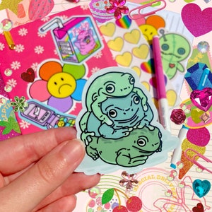 May include: A sticker featuring a cartoon illustration of three green frogs stacked on top of each other. The frogs are on a white background with a light blue outline.
