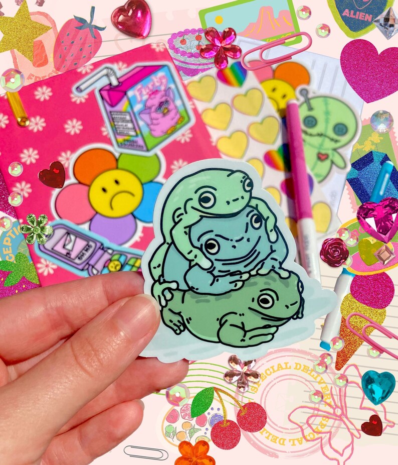 May include: A sticker featuring a cartoon illustration of three green frogs stacked on top of each other. The frogs are on a white background with a light blue outline.