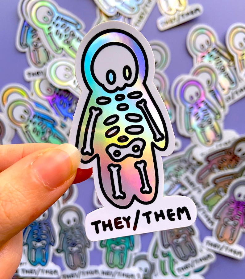 May include: A holographic sticker featuring a skeleton figure with a rainbow gradient. The sticker has a black outline and the text "THEY/THEM" in black lettering.