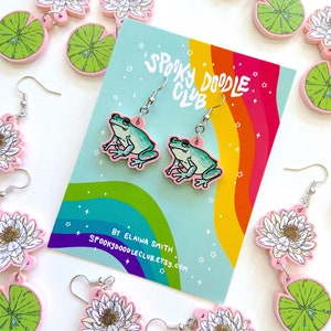 Op de afbeelding: Roze en groene kikker oorbellen met zilveren haken, en bijpassende waterlelie en bloem oorbellen. Een kaart met een regenboog en de tekst "Spooky Doodle Club" is zichtbaar. De oorbellen hebben een grillig, cartoonachtig ontwerp.