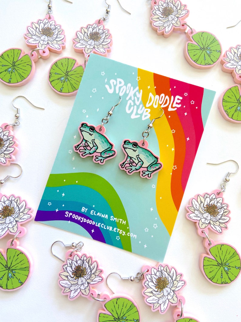 Op de afbeelding: Roze en groene kikker oorbellen met zilveren haken, en bijpassende waterlelie en bloem oorbellen. Een kaart met een regenboog en de tekst "Spooky Doodle Club" is zichtbaar. De oorbellen hebben een grillig, cartoonachtig ontwerp.
