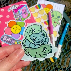 May include: A sticker featuring three green frogs stacked on top of each other. The top frog has its arms around the middle frog, and the middle frog has its arms around the bottom frog. The frogs are all smiling.