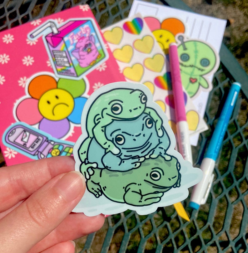 May include: A sticker featuring three green frogs stacked on top of each other. The top frog has its arms around the middle frog, and the middle frog has its arms around the bottom frog. The frogs are all smiling.