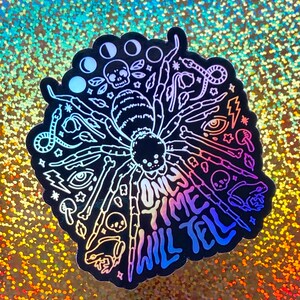 May include: A black and white sticker with a spider design and the text "Only time will tell". The sticker has a holographic finish and features a variety of other designs, including moons, skulls, snakes, and eyes.
