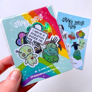 May include: A set of spooky themed enamel pins featuring a voodoo doll, a skeleton, a frog, and a ghost. The pins are packaged in a clear plastic bag with a rainbow background and the text "Spooky Doodle Club".