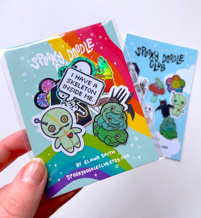 May include: A set of spooky themed enamel pins featuring a voodoo doll, a skeleton, a frog, and a ghost. The pins are packaged in a clear plastic bag with a rainbow background and the text "Spooky Doodle Club".