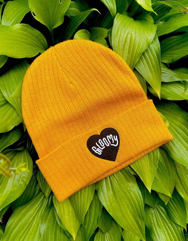 May include: A mustard yellow beanie with a black heart-shaped patch that says "BLOOMY" in white lettering.