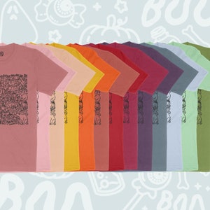 K&ouml;nnte beinhalten: Eine Reihe von 12 T-Shirts in verschiedenen Farben, mit einem schwarz-wei&szlig;en Grafikdruck eines Geistes, K&uuml;rbissen und dem Wort "Boo!" auf der Vorderseite. Die Shirts sind in einem Farbverlauf von Pink bis Gr&uuml;n angeordnet.