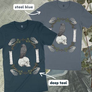 May include: Two t-shirts, one steel blue and one deep teal, featuring a graphic design. The design includes a raven perched on a skull, surrounded by a wreath, candles, and skeletal hands. The shirts are short-sleeved.