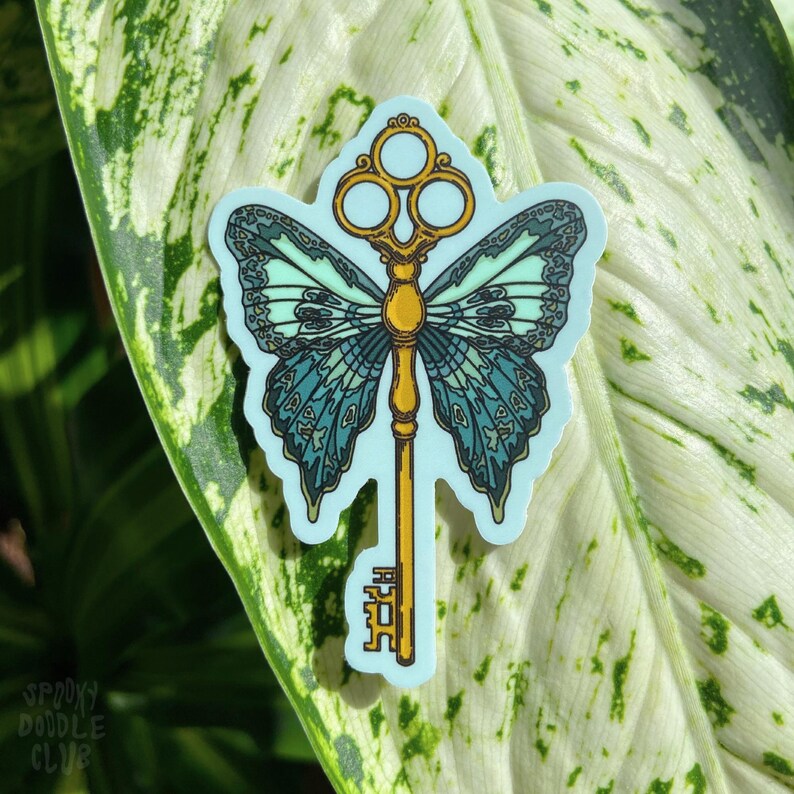 May include: A sticker featuring a gold key with a butterfly design. The butterfly wings are blue and green with a light blue background. The key has a decorative top with three circles.